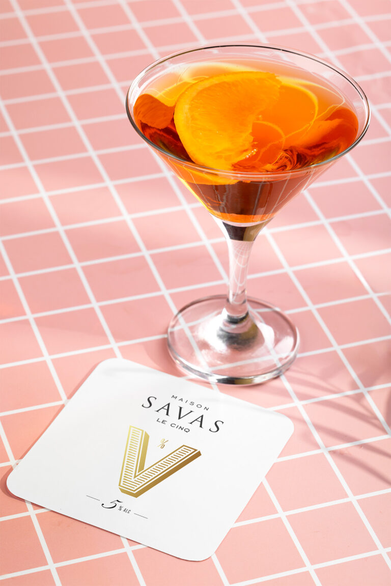 Un cocktail frais à base de vin blanc 5% Le Cinq Maison SAVAS, sur fond rose estival avec sous-verre doré.
